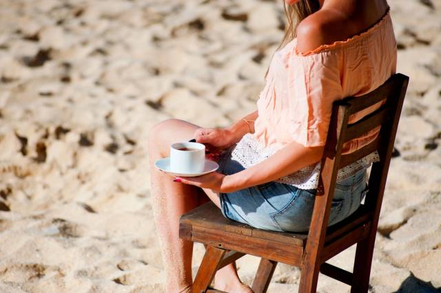 Une femme se détend avec un café sur une plage ensoleillée, assise sur une chaise en bois