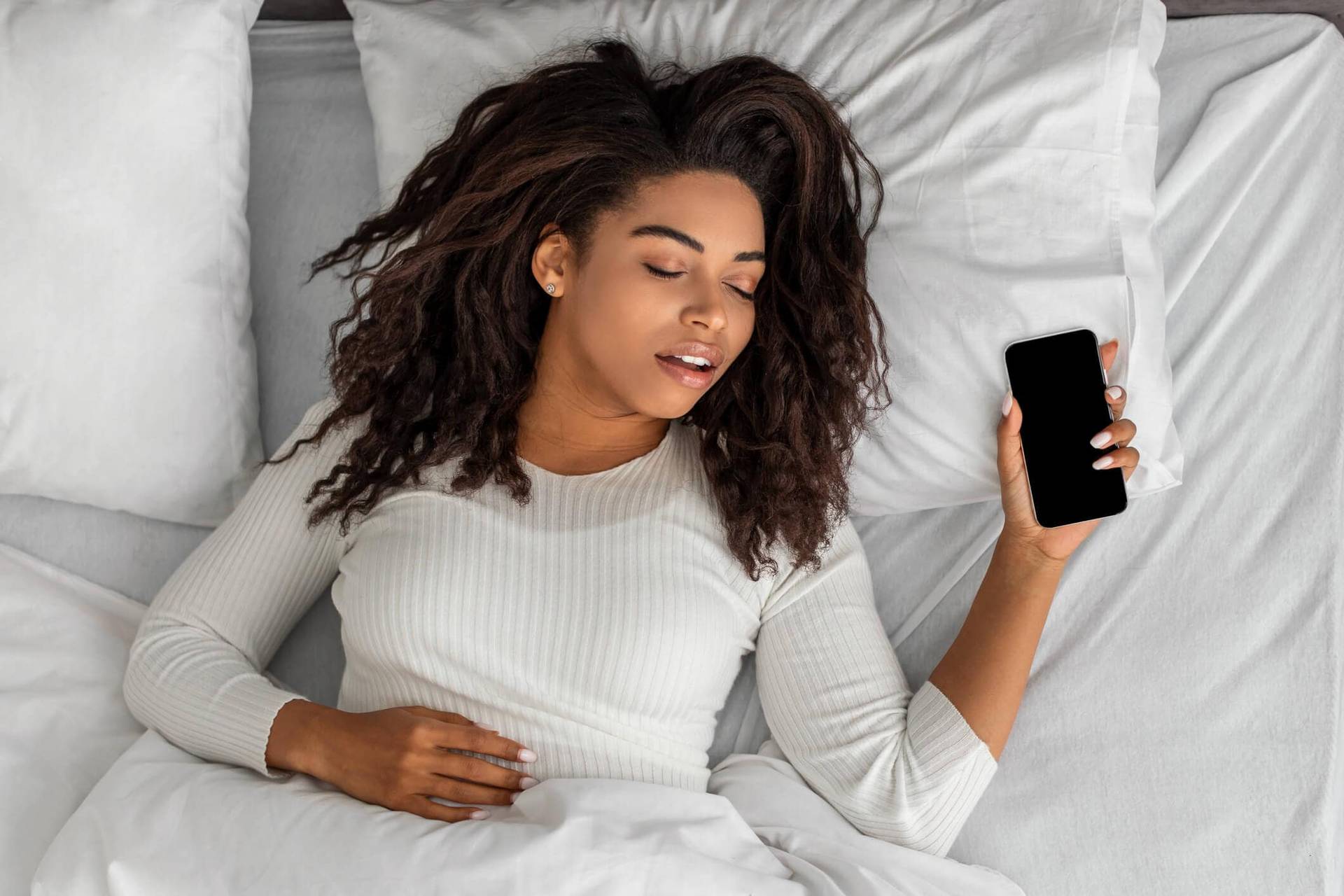 Femme allongée sur un lit, les yeux fermés, tenant un téléphone.