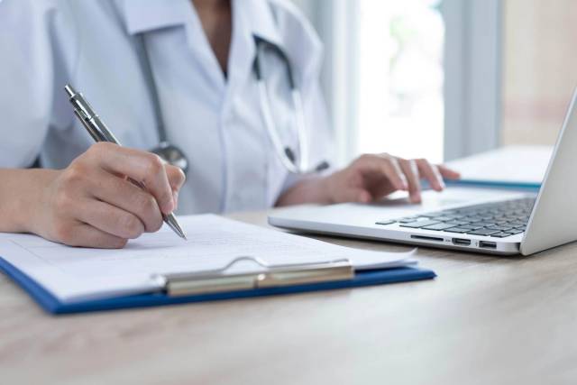 Professionnel de santé écrivant sur un document près d’un ordinateur.
