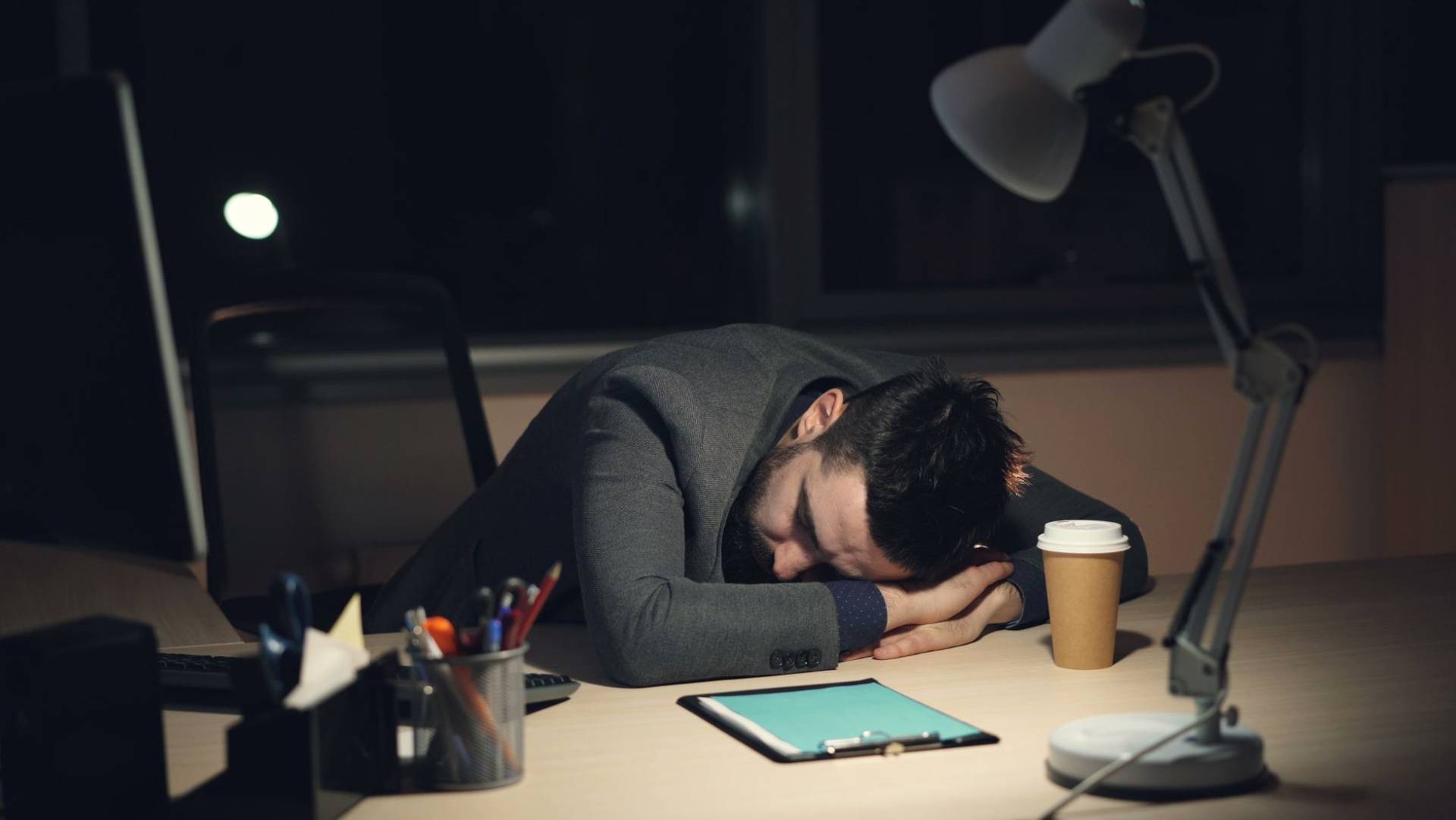 Homme en costume de bureau dormant sur son bureau avec du café et une tablette à proximité, symbolisant la fatigue