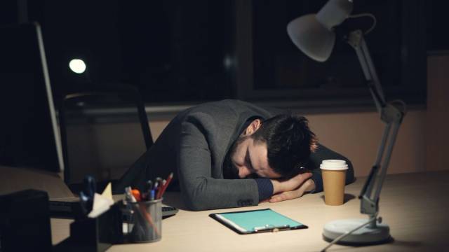 Homme en costume de bureau dormant sur son bureau avec du café et une tablette à proximité, symbolisant la fatigue