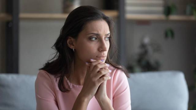 Femme pensive assise sur un canapé, mains jointes près du visage.