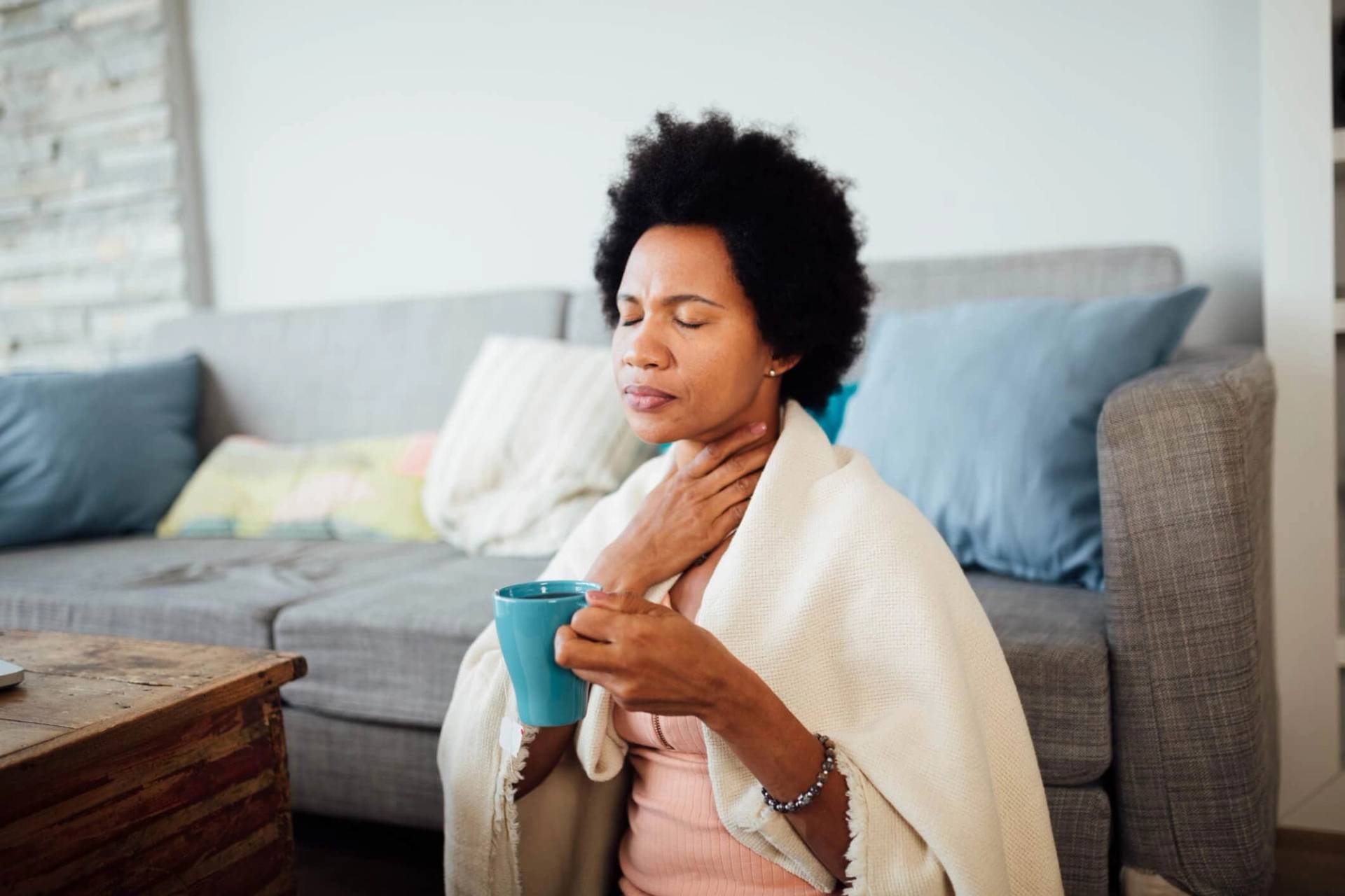 Femme tenant sa gorge et une tasse, sous une couverture.