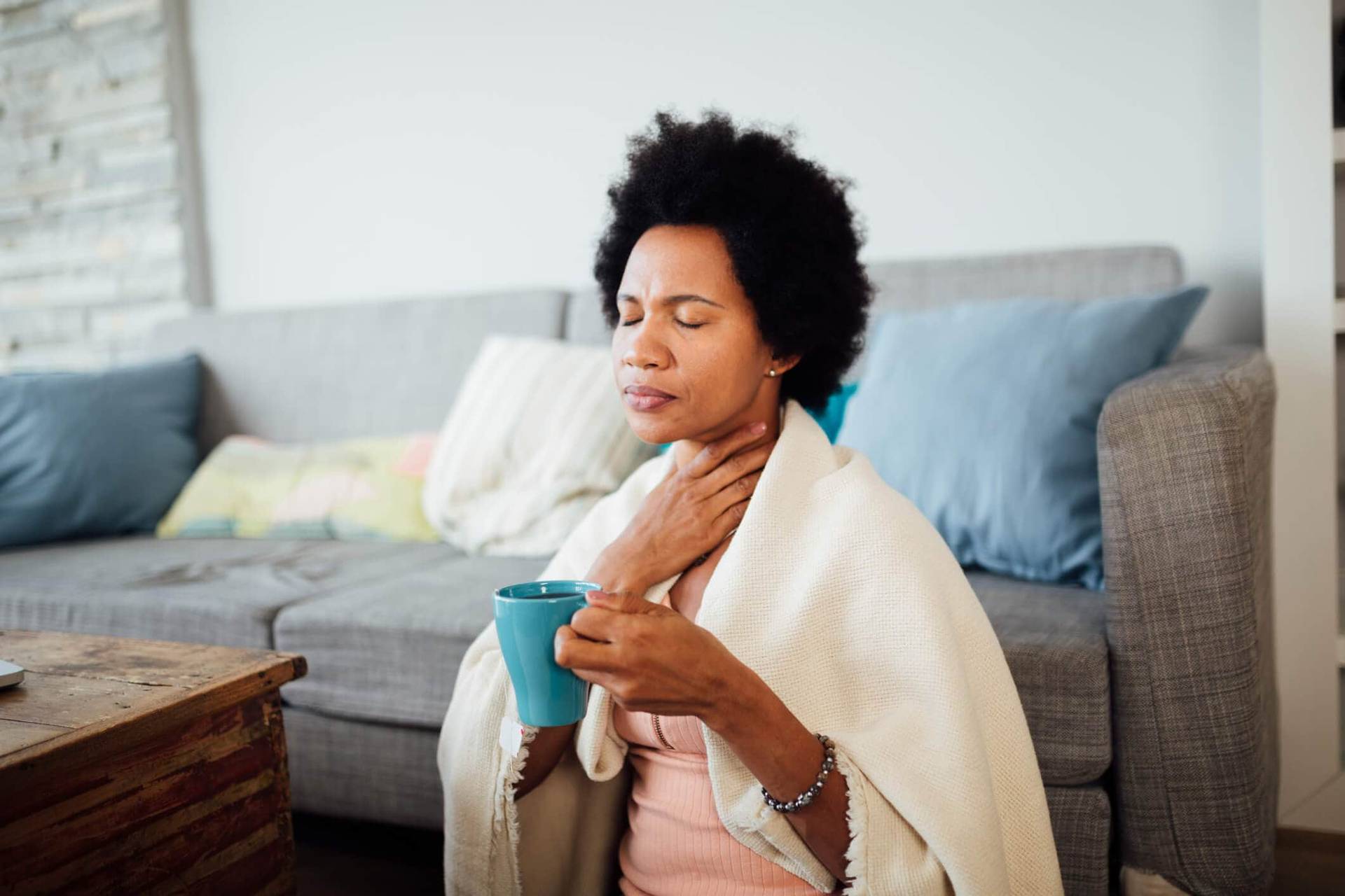 Femme tenant sa gorge et une tasse, sous une couverture.