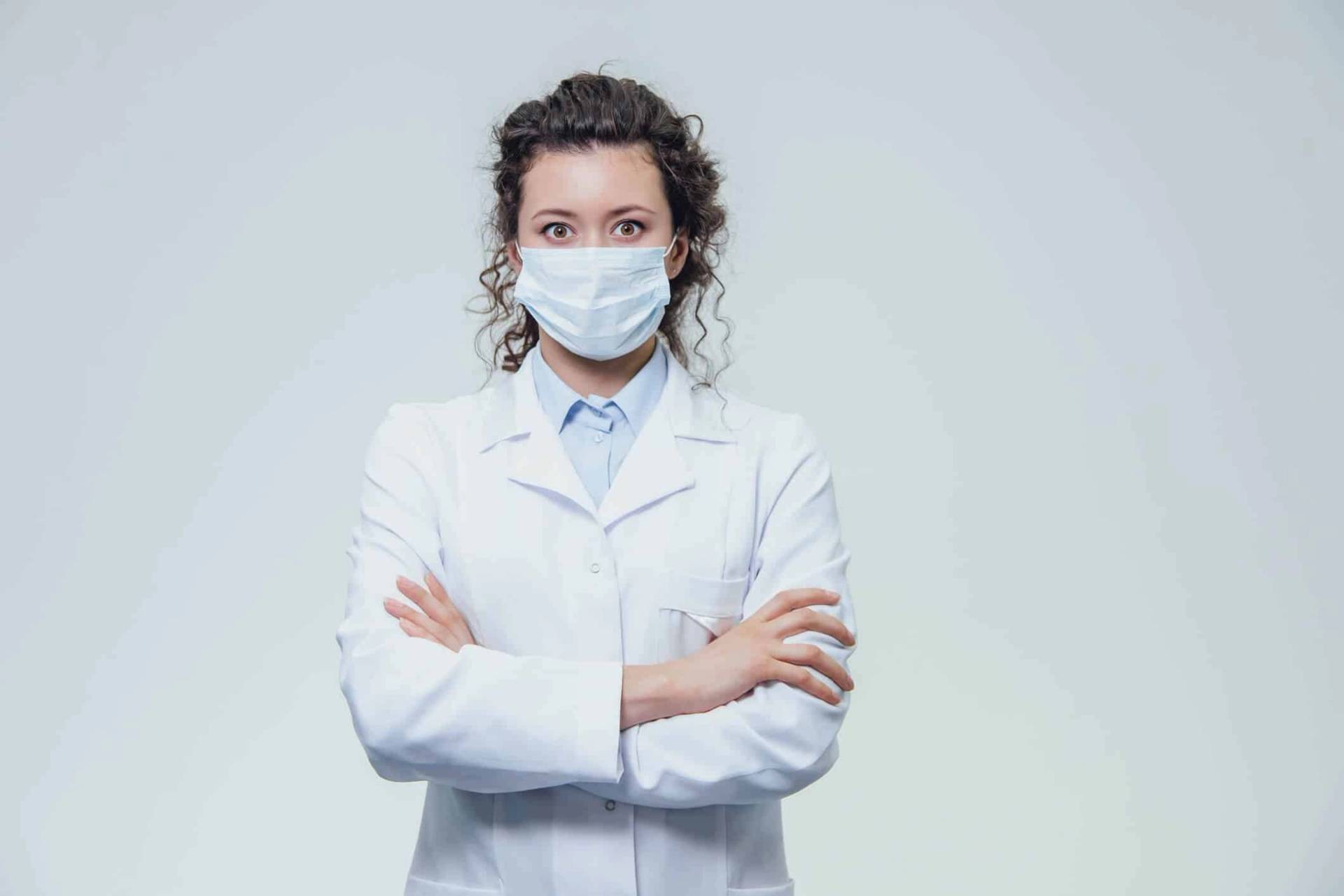 Femme en blouse et masque chirurgical, les bras croisés.