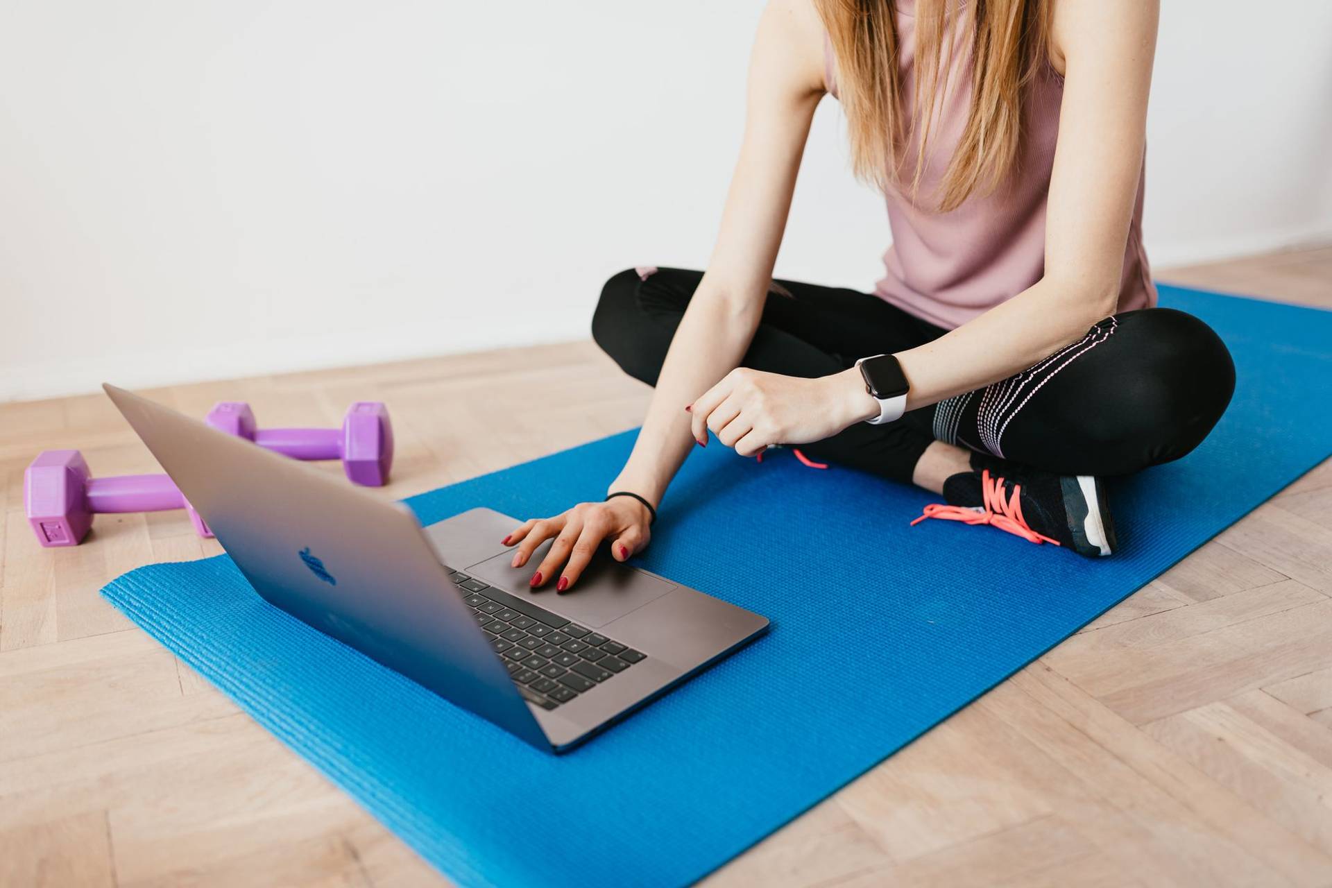 Personne assise sur un tapis de sport devant un ordinateur portable, avec des haltères à proximité.