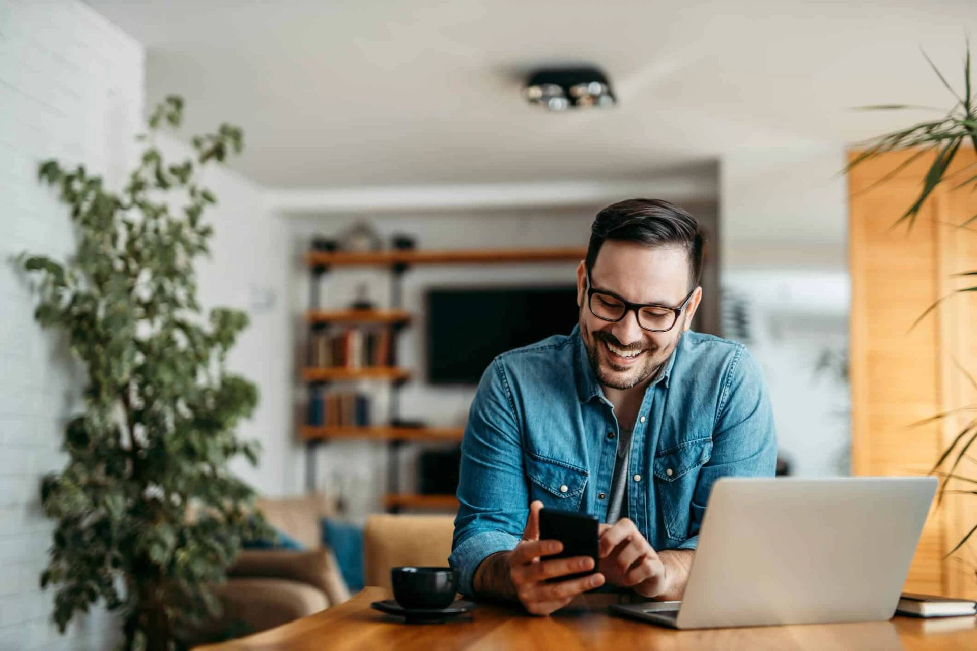 Homme souriant regardant son téléphone près d’un ordinateur.