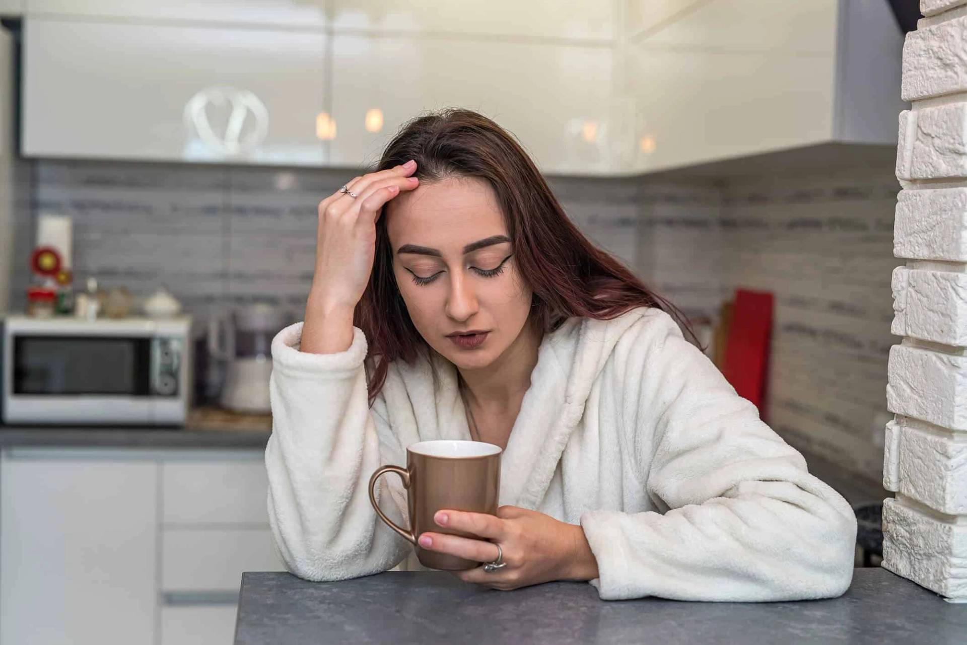 Une femme en peignoir blanc assise dans une cuisine, l'air fatigué, qui tient une tasse marron et se touche le front.