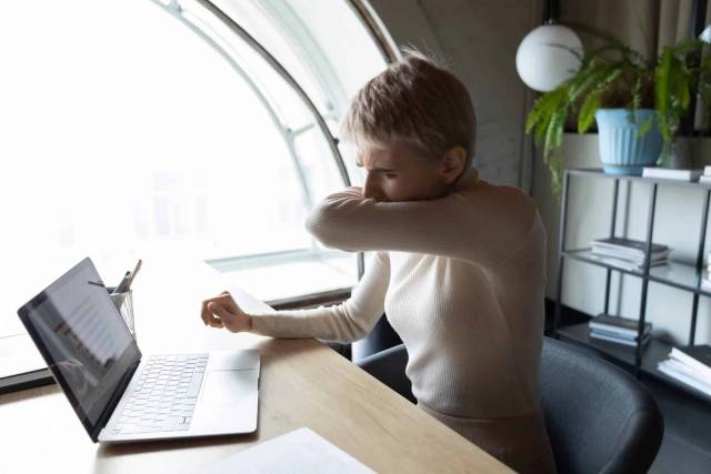 Femme toussant dans son coude à son bureau.