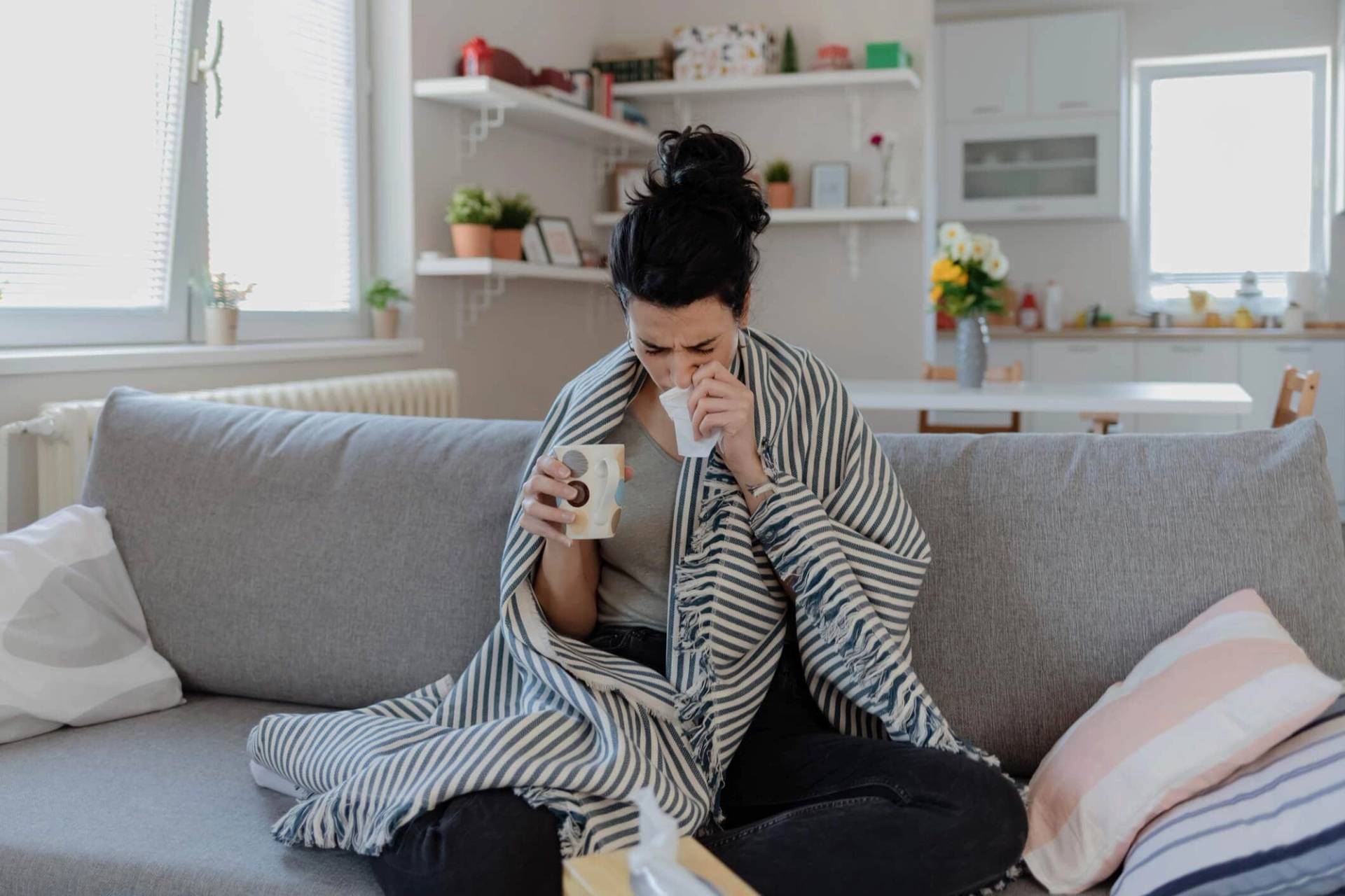 Femme sur un canapé avec couverture, tasse et mouchoir.