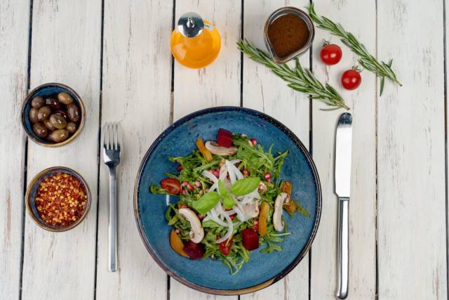 Salade de légumes dans une assiette bleue, accompagnée d’olives et d’huile sur une table en bois.