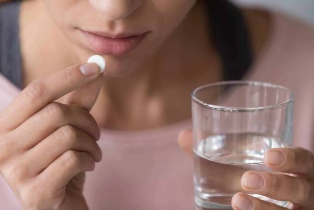 Femme sur le point d’avaler un comprimé blanc, un verre d’eau à la main.