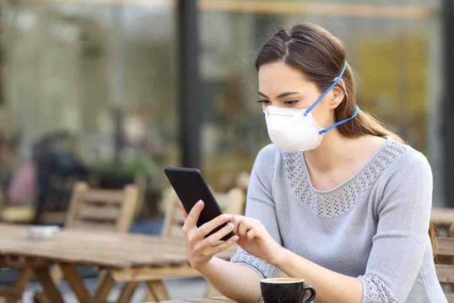 Une femme portant un masque consulte son téléphone en terrasse.