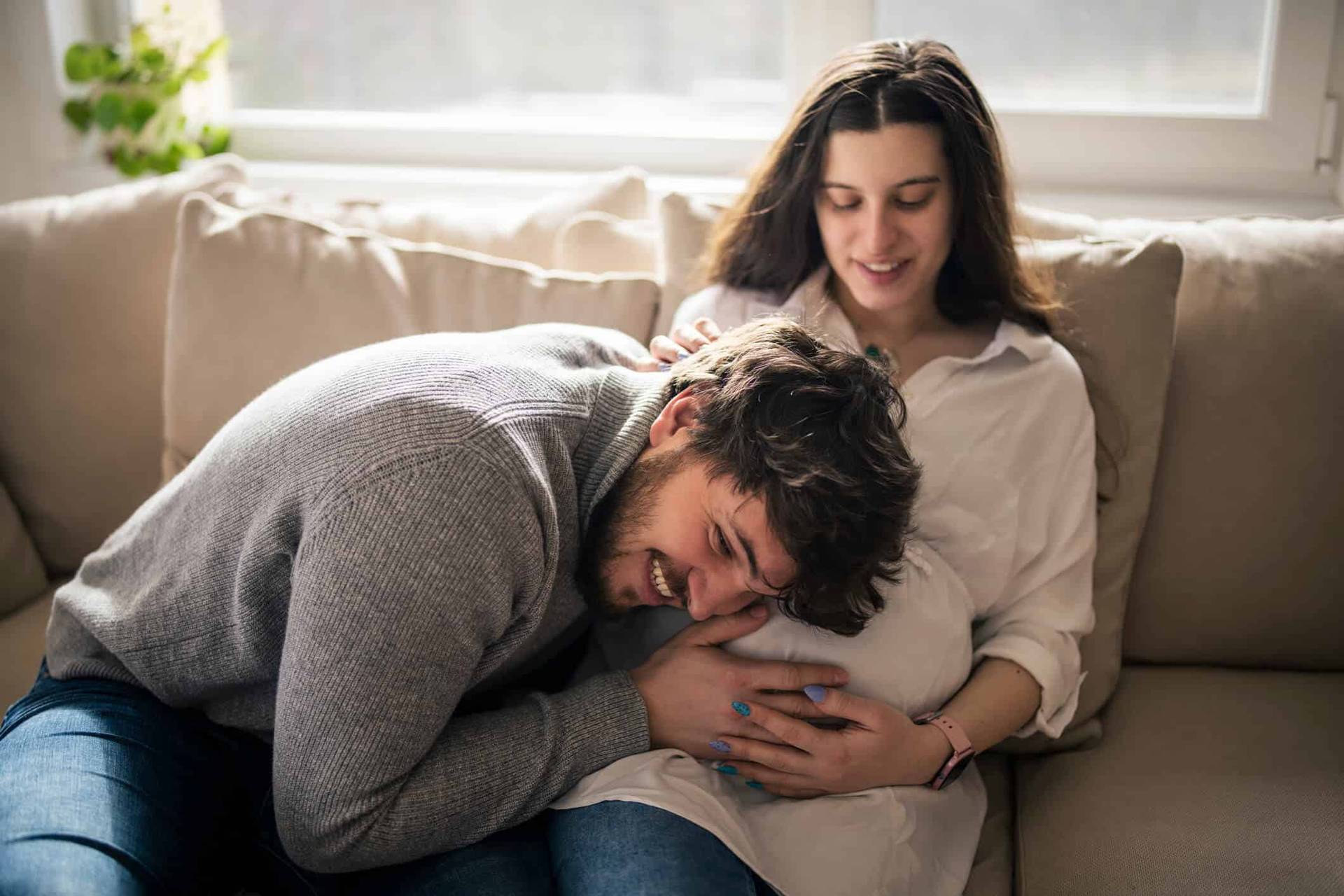 Un homme pose la tête sur le ventre arrondi d’une femme assise sur un canapé.