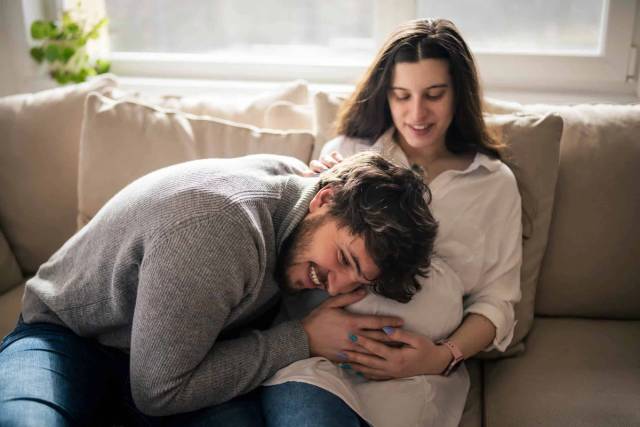 Un homme pose la tête sur le ventre arrondi d’une femme assise sur un canapé.