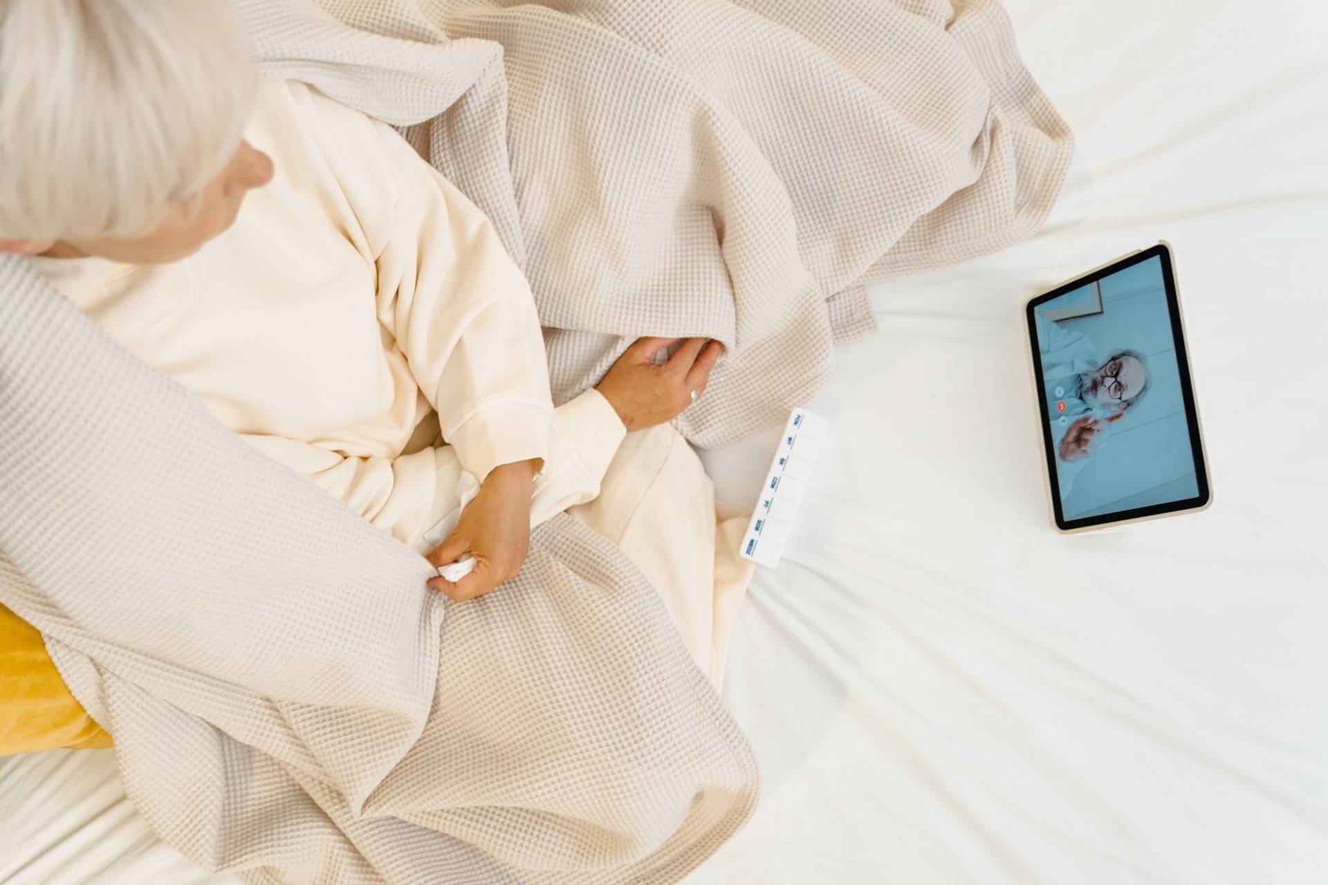 Femme âgée alitée en téléconsultation avec un médecin sur une tablette.