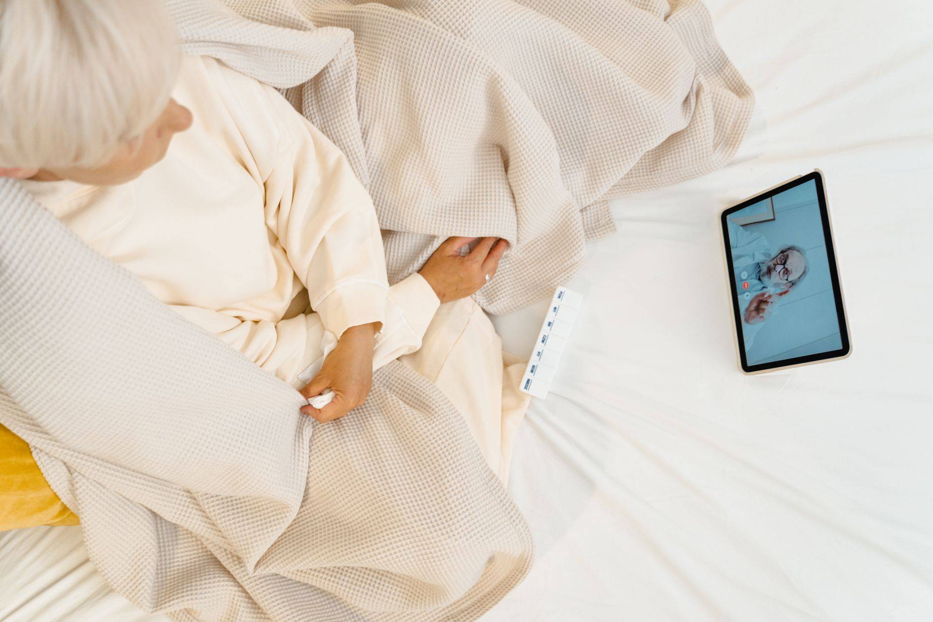 Femme âgée alitée en téléconsultation avec un médecin sur une tablette.