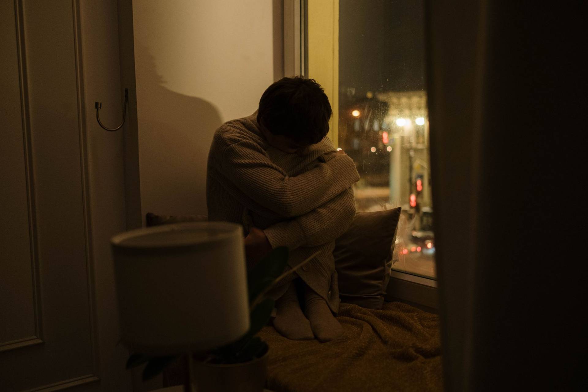 Une femme assise seule près d’une fenêtre, la nuit, la tête posée sur ses genoux.