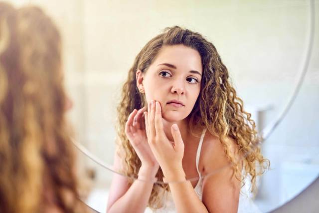 Une femme examine la peau de sa joue dans un miroir.