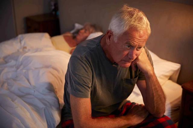 Un homme âgé assis au bord du lit paraît préoccupé.