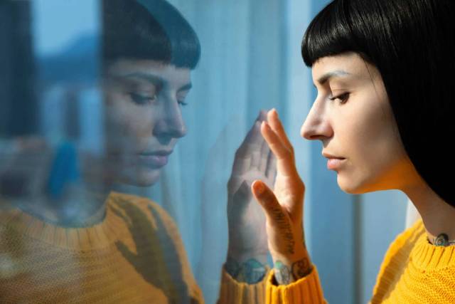 Une femme regarde à travers une fenêtre, une main posée sur la vitre.