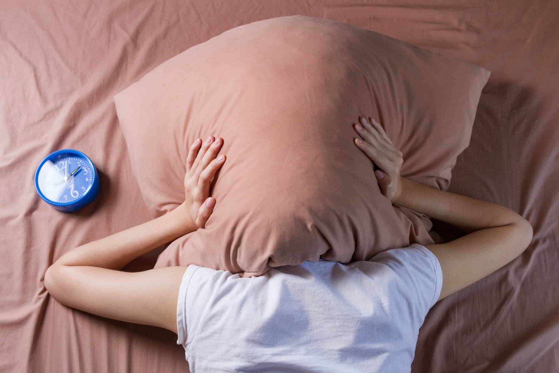 Personne allongée sur son lit, la tête cachée sous un oreiller, à côté d’un réveil.