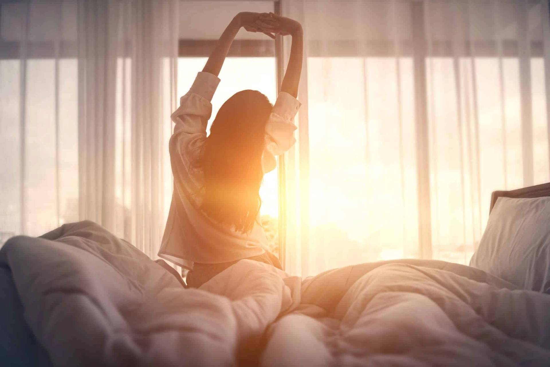 Personne assise sur un lit, s’étirant devant une fenêtre ensoleillée.