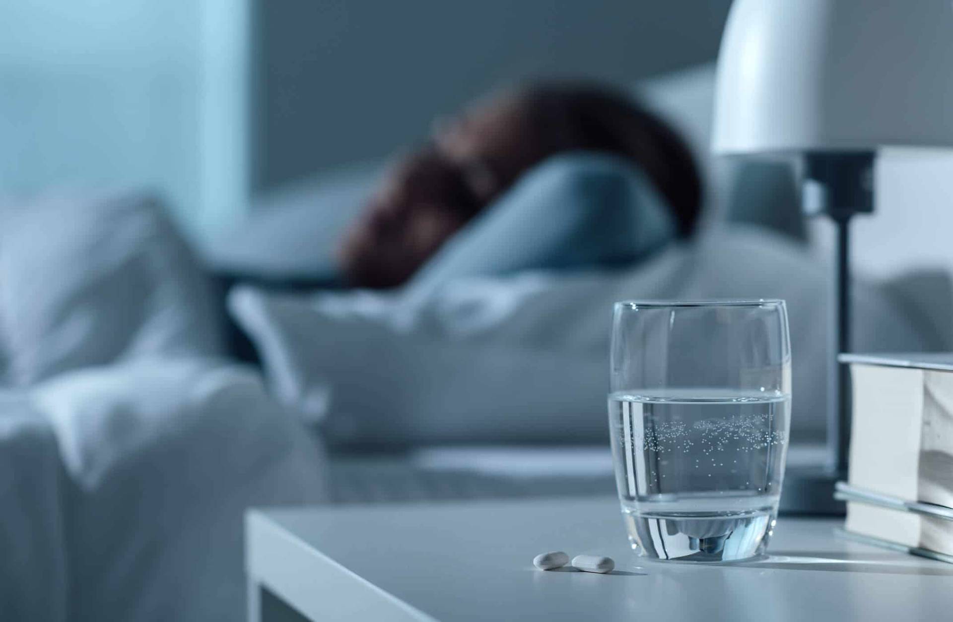 Verre d’eau et comprimés sur une table de chevet, personne allongée en arrière-plan.