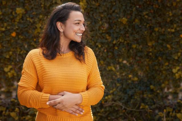 Une femme enceinte en pull jaune pose les mains sur son ventre en extérieur.