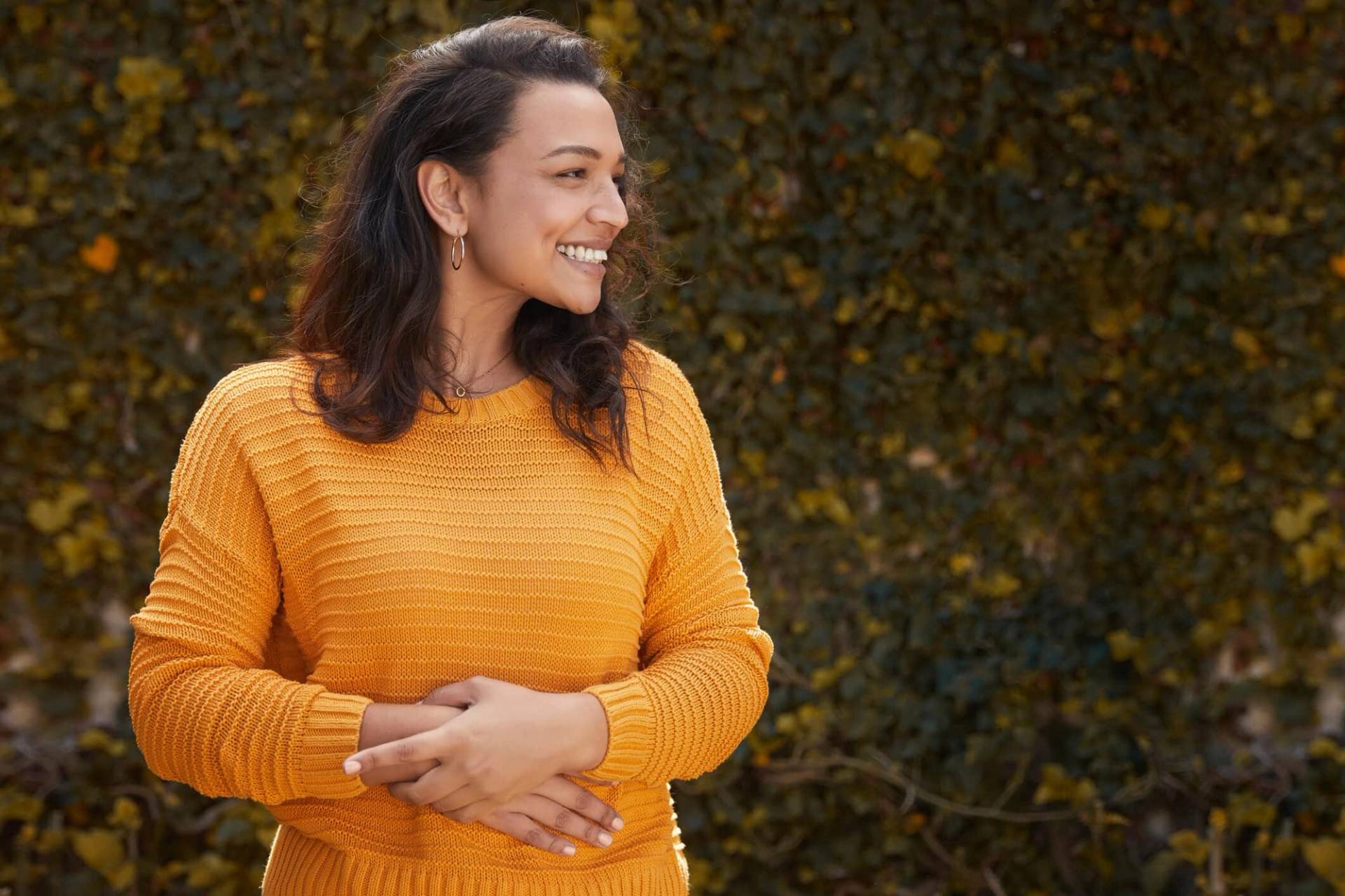 Une femme enceinte en pull jaune pose les mains sur son ventre en extérieur.