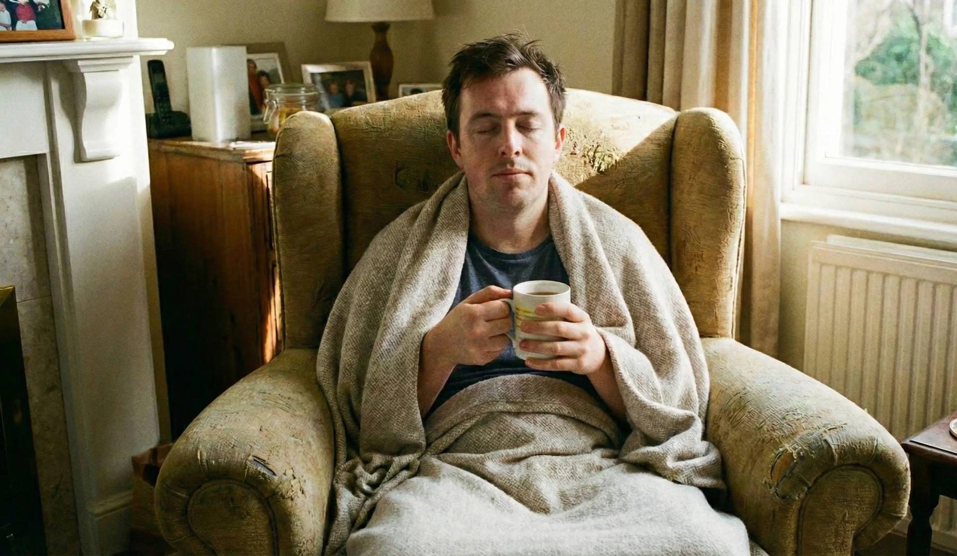 Un homme aux yeux fermés, enveloppé dans une couverture en tricot beige et tenant une tasse, se repose paisiblement dans