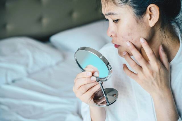 Une femme examine les boutons de son visage dans un miroir à main.