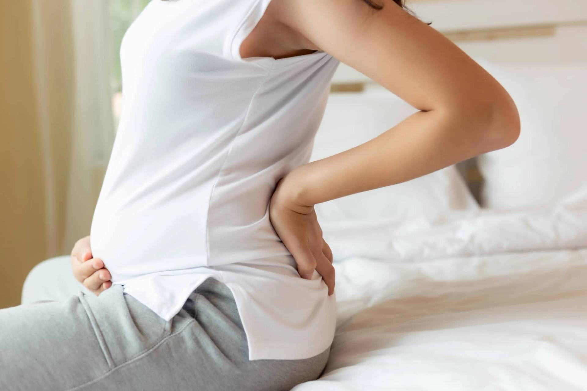 Femme enceinte assise sur un lit, une main sur le ventre et l’autre au bas du dos.