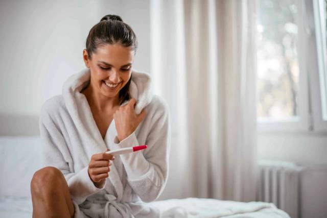 Femme souriante tenant un test de grossesse sur un lit.