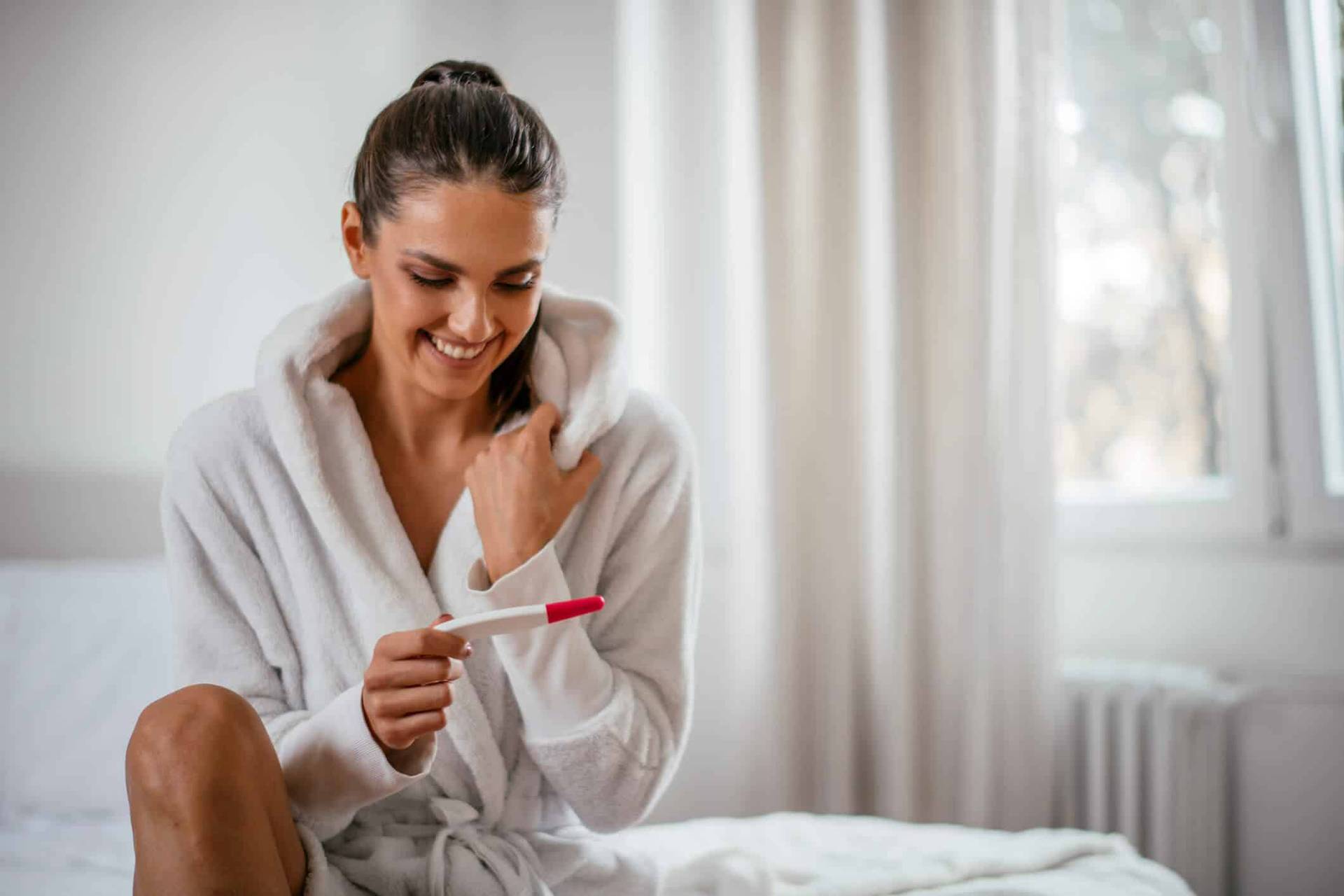 Femme souriante tenant un test de grossesse sur un lit.
