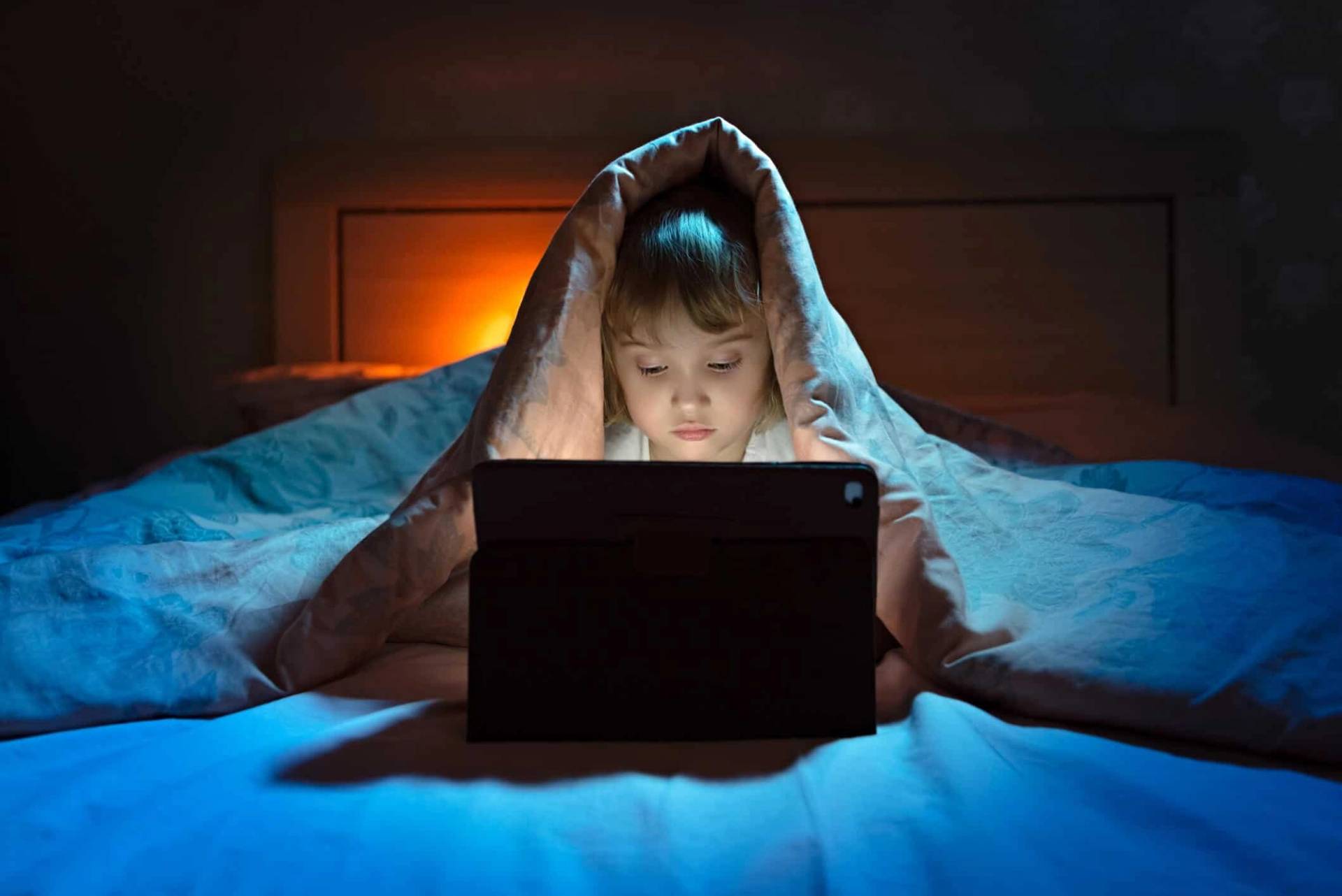 Enfant sous une couverture utilisant une tablette dans son lit.