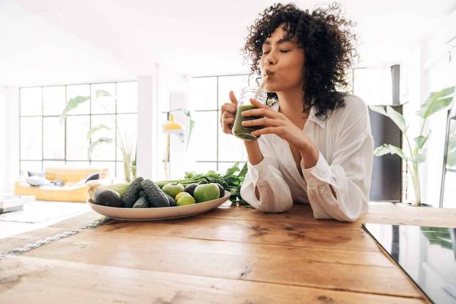 Femme buvant un jus vert dans une cuisine avec des légumes.