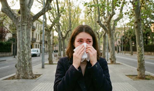 Femme souffrant d'allergie aux platanes éternuant dans un mouchoir sur une rue bordée d'arbres.