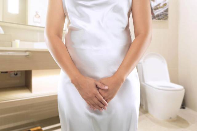 Femme debout devant des toilettes, les mains devant l’entrejambe.