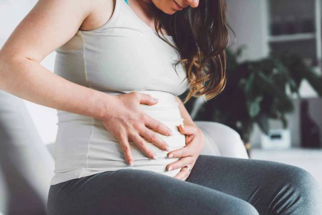 Mains posées sur le ventre d’une personne enceinte en tee-shirt gris.