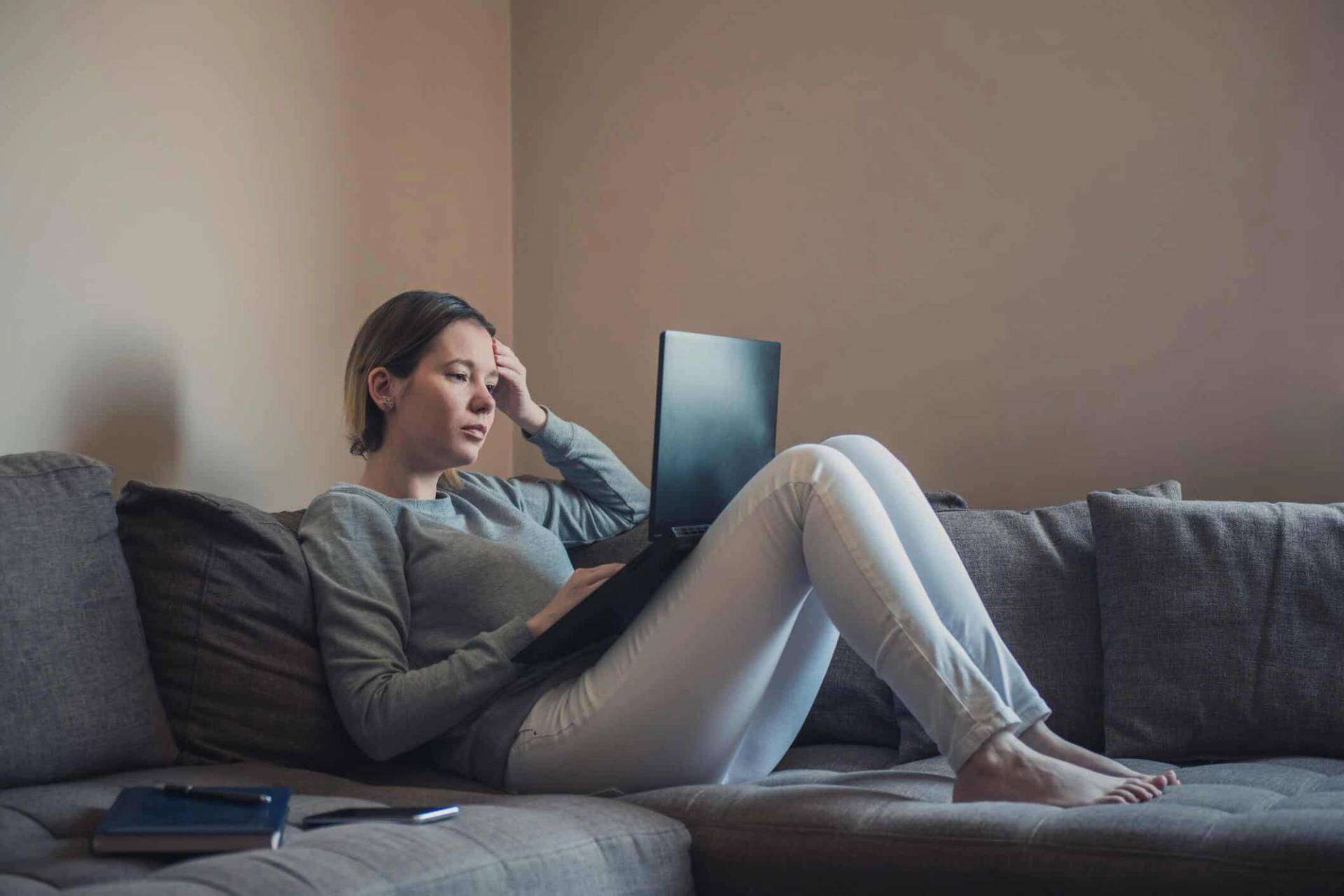 Femme allongée sur un canapé avec un ordinateur, main sur le front.