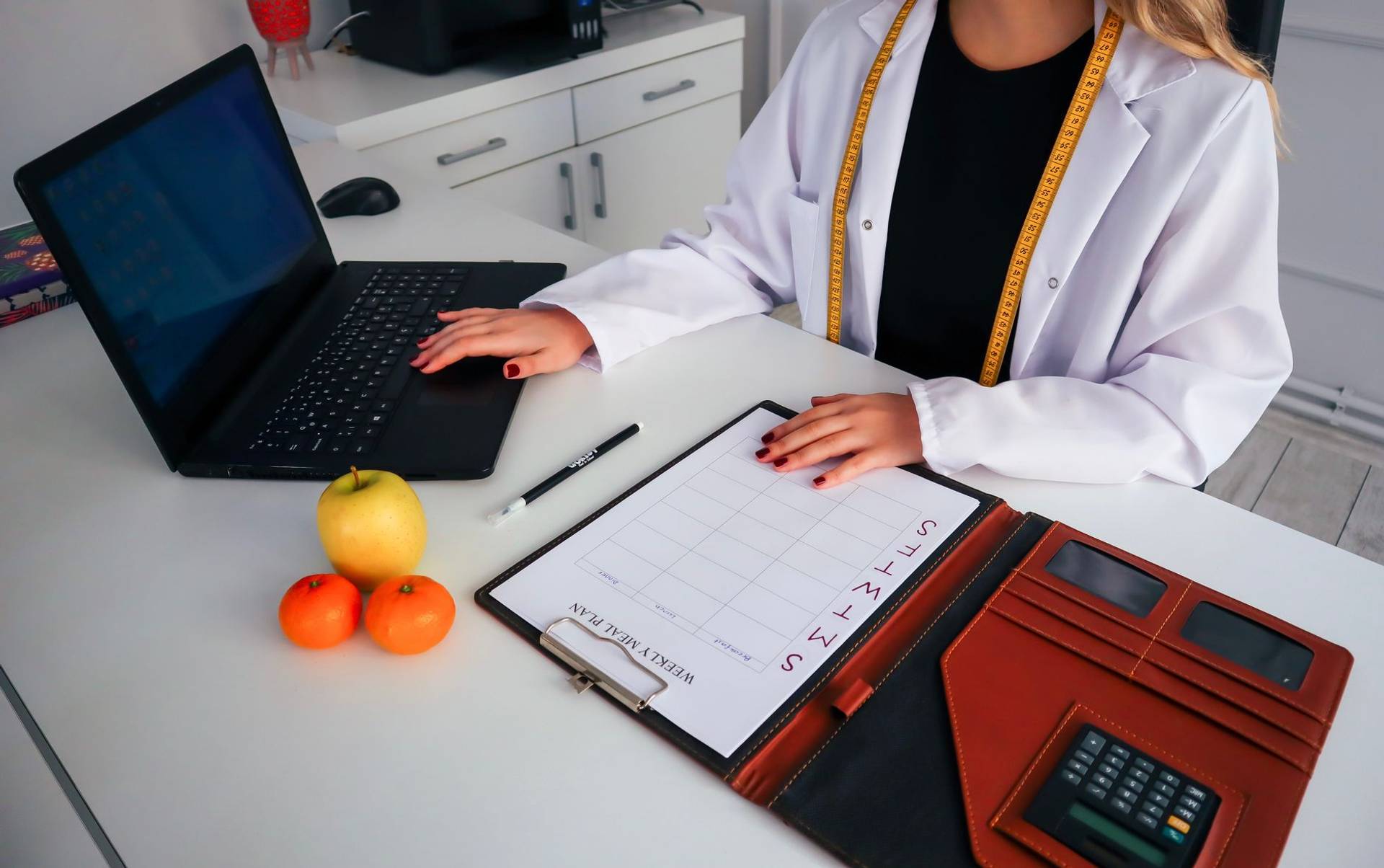 Professionnelle en blouse devant un ordinateur et un planning de repas, avec des fruits sur le bureau.
