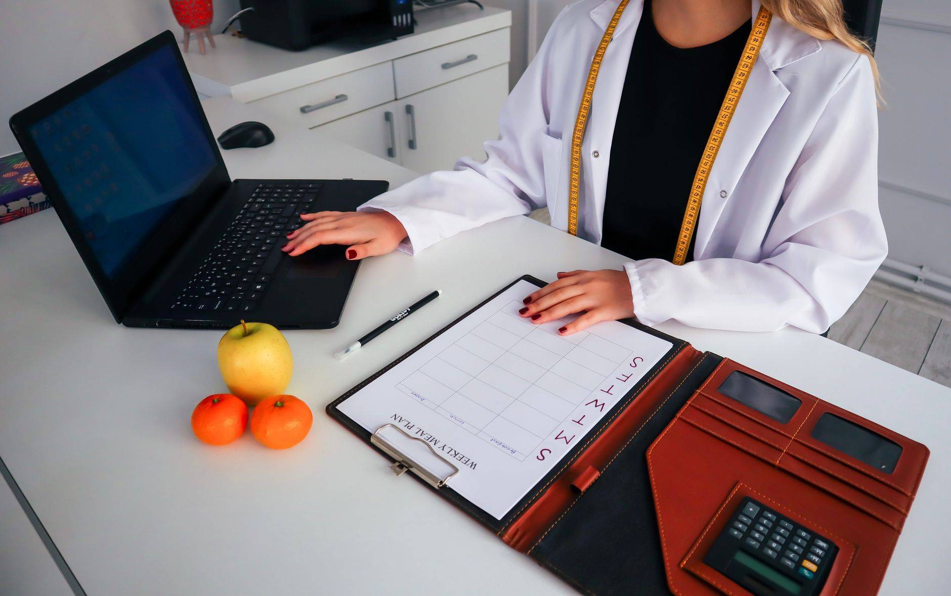 Professionnelle en blouse devant un ordinateur et un planning de repas, avec des fruits sur le bureau.