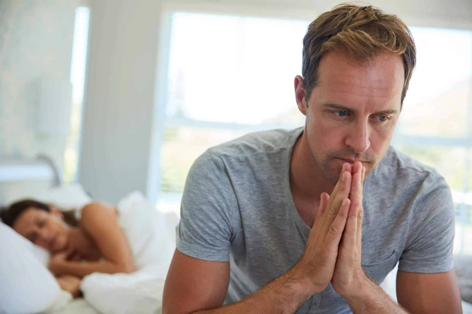 Homme assis au bord du lit, une femme allongée derrière lui.