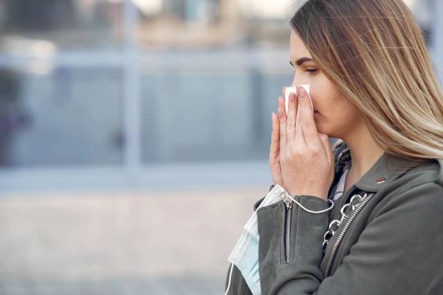 Une femme éternue en plein air tout en tenant un mouchoir et un masque, reflétant des préoccupations de santé