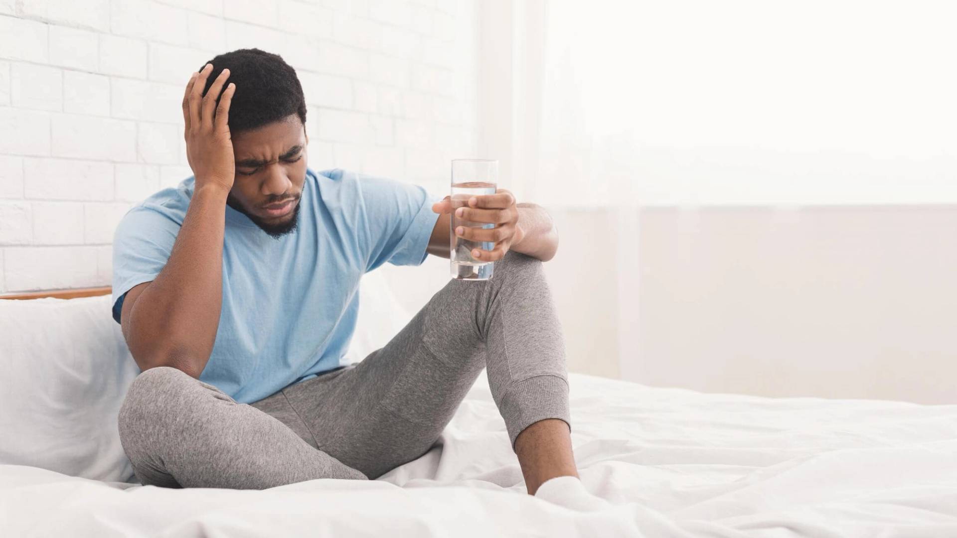 Homme assis sur un lit, une main sur la tête et un verre d’eau dans l’autre.