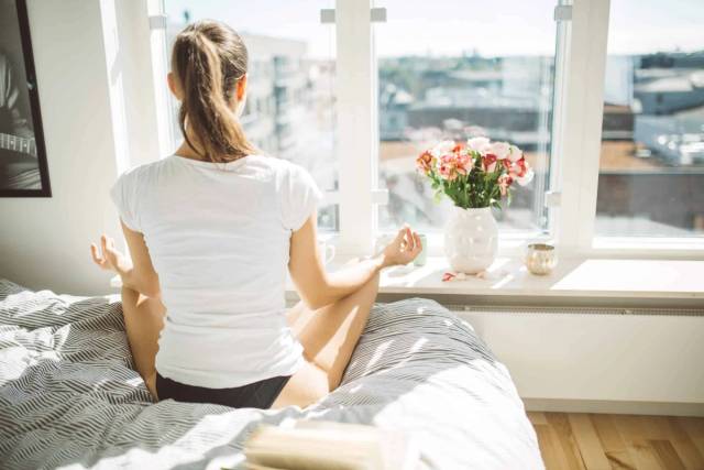 Femme assise en méditation sur un lit près d’une fenêtre.