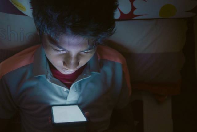 Garçon allongé sur le lit, regardant intensément un écran de smartphone lumineux