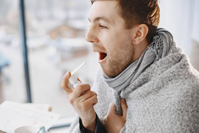 Un homme adulte utilise un spray pour la gorge, enveloppé dans une couverture, ayant l'air mal à l'aise à