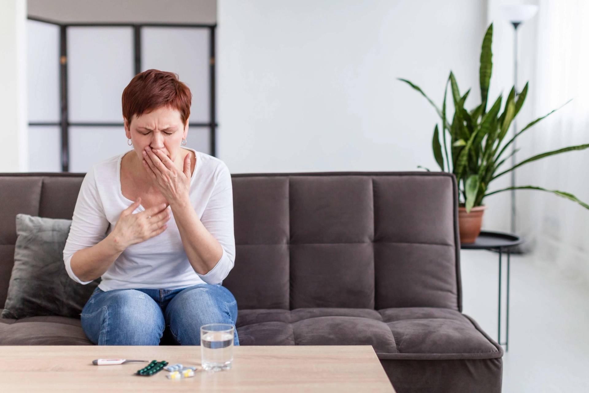 Femme assise sur un canapé, se tenant la poitrine et toussant, avec des médicaments posés sur la table devant elle